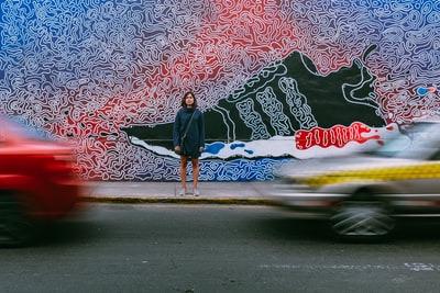 During the day, wearing a blue jacket women stood on the cement and the two cars on the road car time-lapse photography
